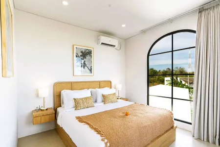 Bright tropical bedroom with wooden bed, orange throw blanket, and ocean view through arched black-framed window. - Bali Villas